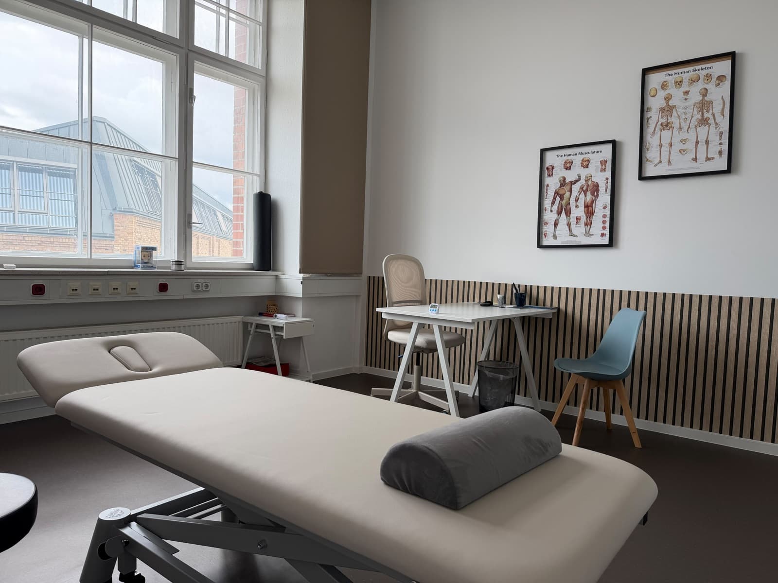 Behandlungsraum mit Therapieliege, Schreibtisch und Anatomie-Postern